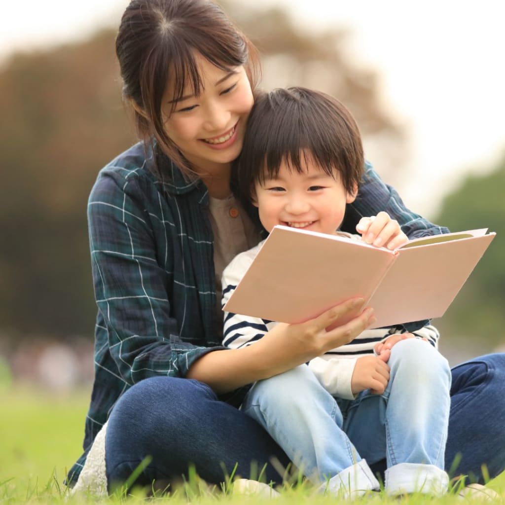 A woman and child sit on grass, smiling while reading a book together.